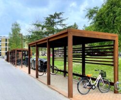 Casa 44 en modulopbygget cykeloverdækning i cortenstål med installeret cykelstativer og beklædning af mahogni planker, som står ved siden af en stor lejlighedsbygning i Vollsmose