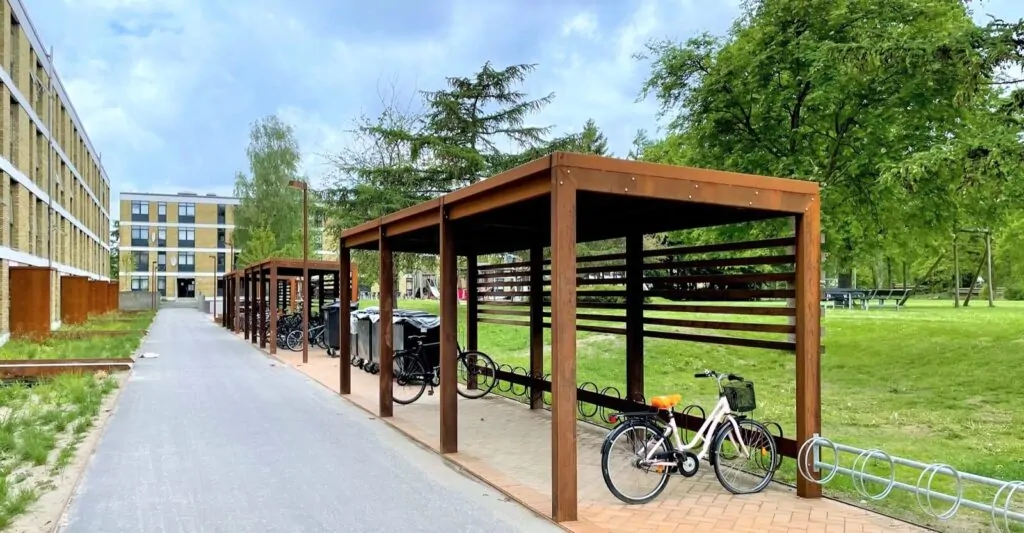 en modulopbygget cykeloverdækning i cortenstål med installeret cykelstativer og beklædning af mahogni planker, som står ved siden af en stor lejlighedsbygning i Vollsmose